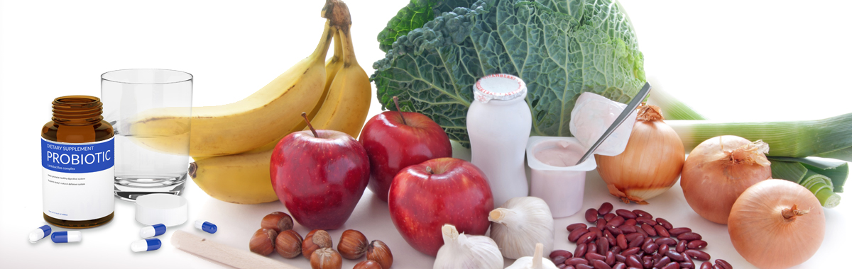 Bananas, apples, onions, yogurt, and various healthy foods along with a bottle of probiotics and a glass of water on a white surface