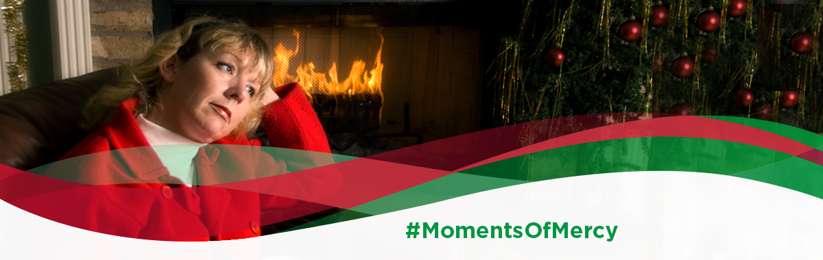 Woman in red sweater and white turtleneck holds head in hand as she sits by the fire dealing with grief