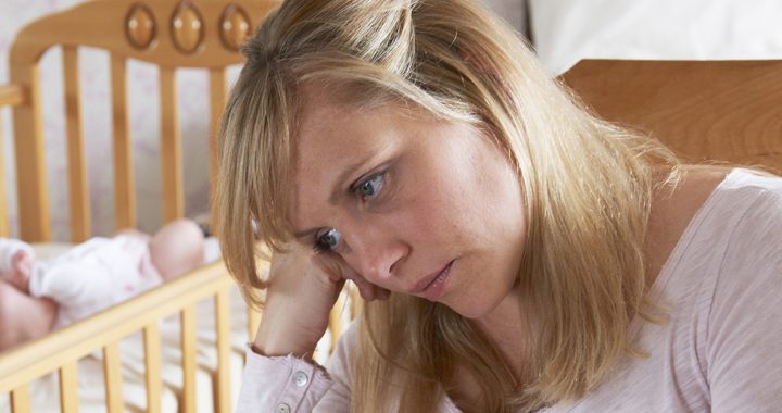 Woman with postpartem depression rests head on hand with fussy baby in the background lying in their crib