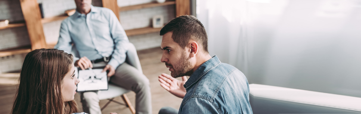 Couple sitting on couch with intense faces looks to be arguiing in front of therapist during marriage counseling