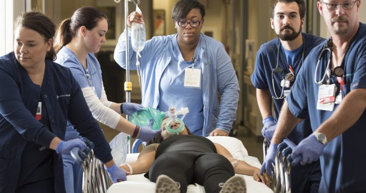 A team of five nurses in blue scrubs attend to a female patient who is unresponsive and laying in a hospital bed