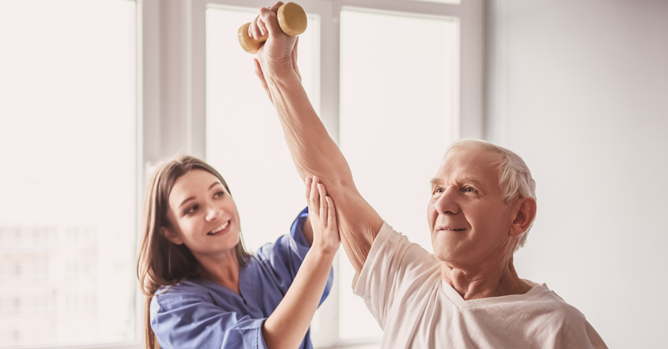 A young female physical therapist helps an older man lift his arm above his head with a small weight in his hand