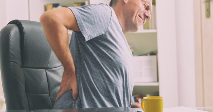 A middle-aged man holds his back in pain as he stands up out of a black office chair with a grimace on his face