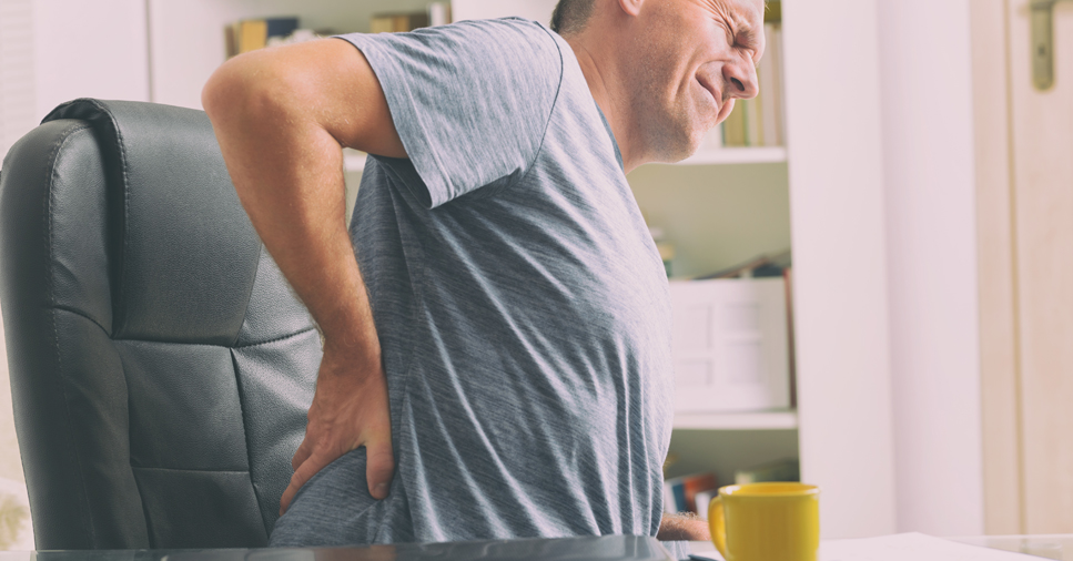 A middle-aged man holds his back in pain as he stands up out of a black office chair with a grimace on his face