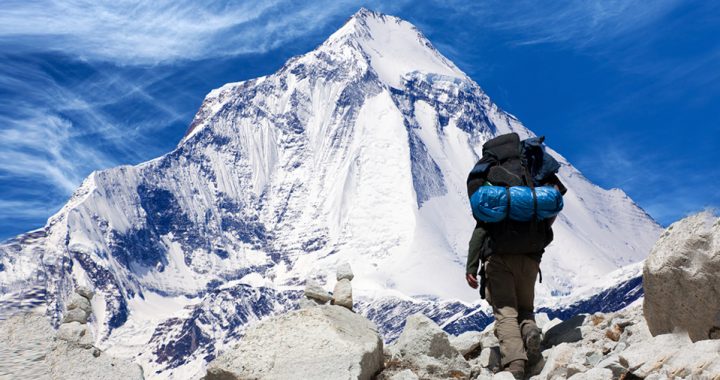 A man with a hiking backpack stands beneath a snow covered mountain