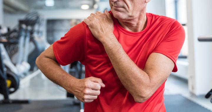 A middle-aged man in a red shirt at the gym clutches his right shoulder with his left hand