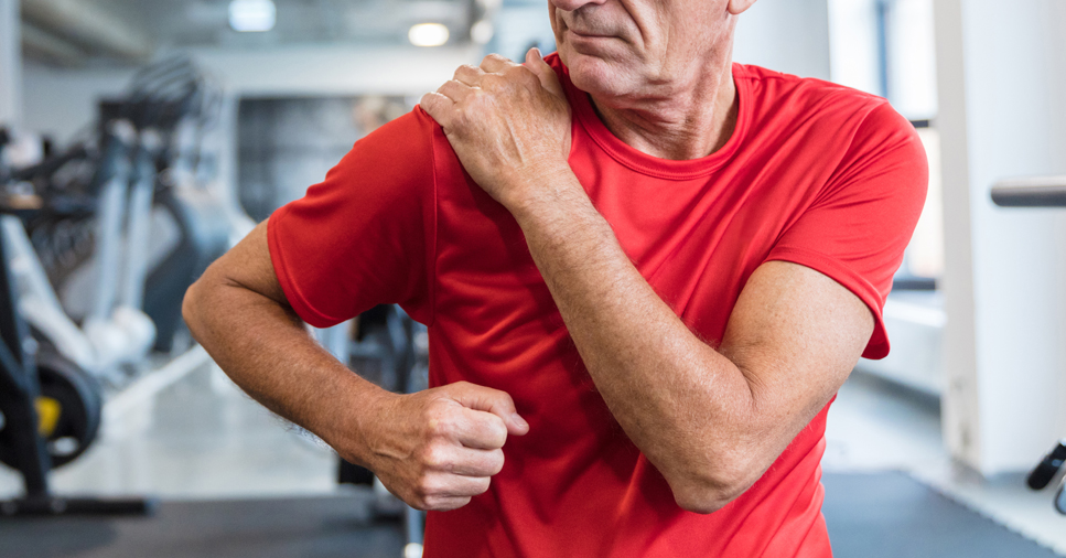 A middle-aged man in a red shirt at the gym clutches his right shoulder with his left hand