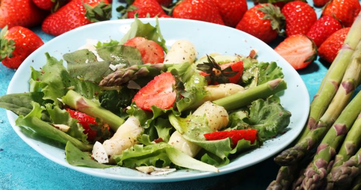 A salad with spring vegetables and fruits such as strawberries and asparagus on a white plate sits on a table