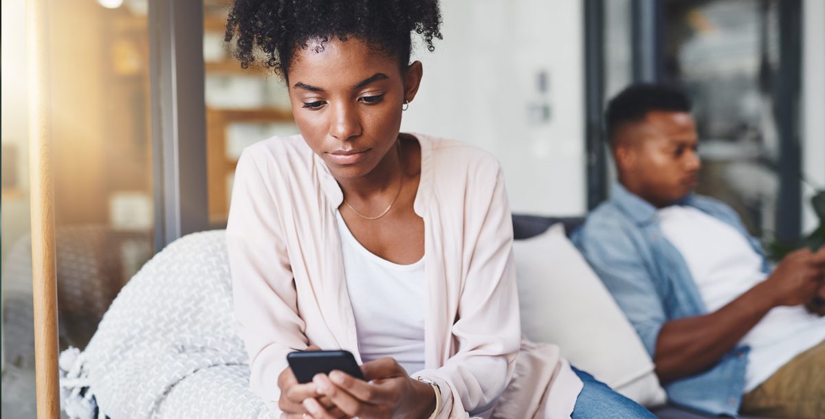 A woman on her phone browsing social media sits facing away from her boyfriend who is also on his phone