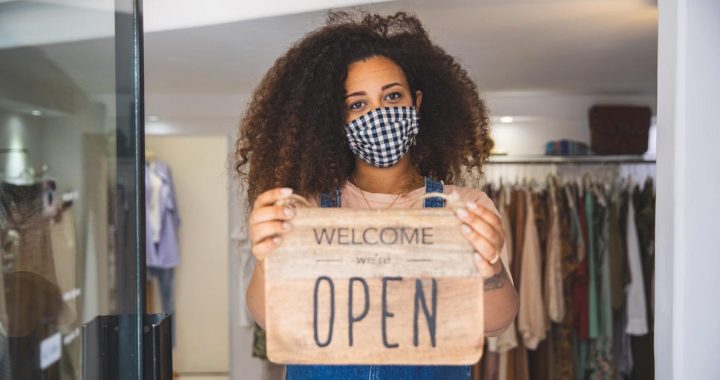 A woman holding a "we are open" sign in front of her store while wearing a face mask.